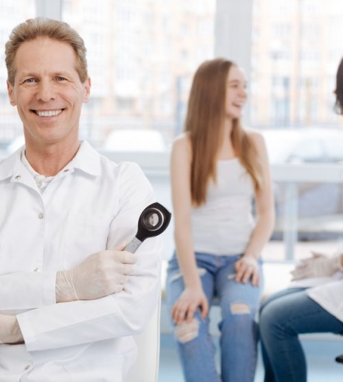 cheerful-qualified-handsome-dermatologist-working-in-the-clinic-and-holding-dermatoscope-while-colleague-talking-to-young-patient-in-the-background