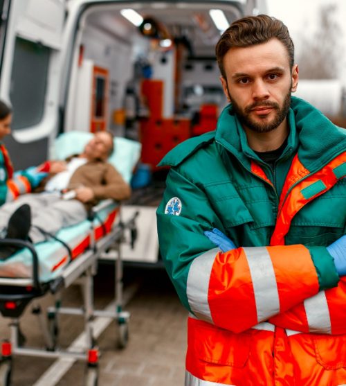 male-paramedic-in-uniform-stands-with-his-arms-crossed-in-front-of-an-ambulance-and-his-colleague-standing-near-patient-s-gurney